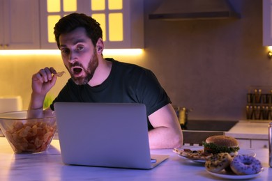 Photo of Man eating chips while using laptop in kitchen at night. Bad habit