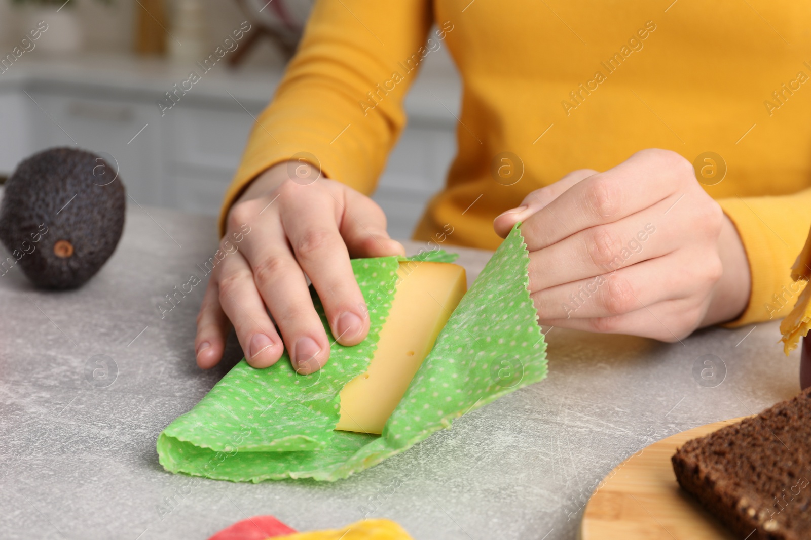 Photo of Woman packing piece of cheese into beeswax food wrap at light table in kitchen, closeup