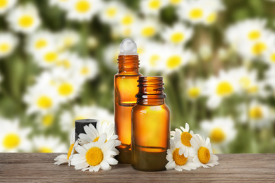 Bottles of essential oil and chamomile flowers on wooden table against blurred background
