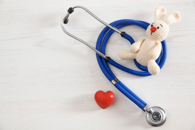 Toy bunny, stethoscope, heart and space for text on white wooden background, top view. Children's doctor