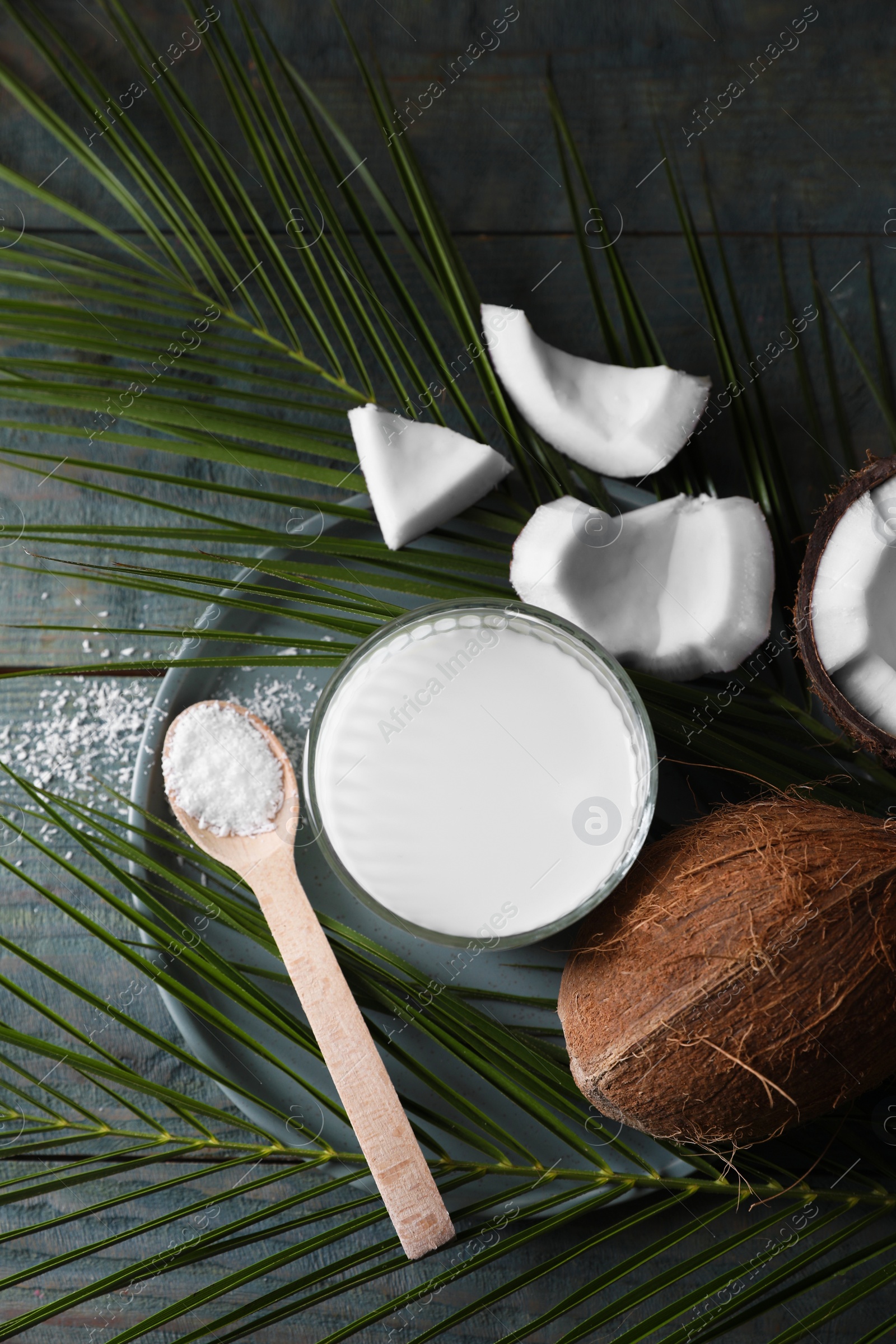 Photo of Glass of delicious coconut milk, spoon with flakes, nuts and palm leaves on wooden table, flat lay