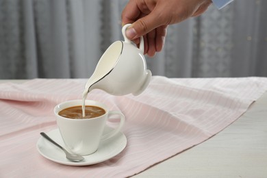 Woman pouring milk into cup with coffee at table indoors, closeup