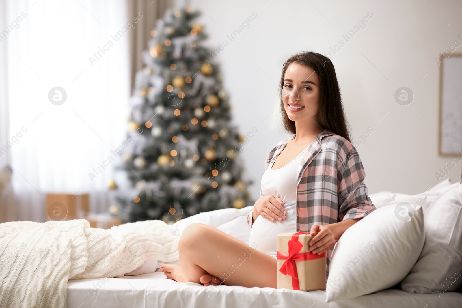 Photo of Happy pregnant woman with Christmas gift box on bed at home. Expecting baby