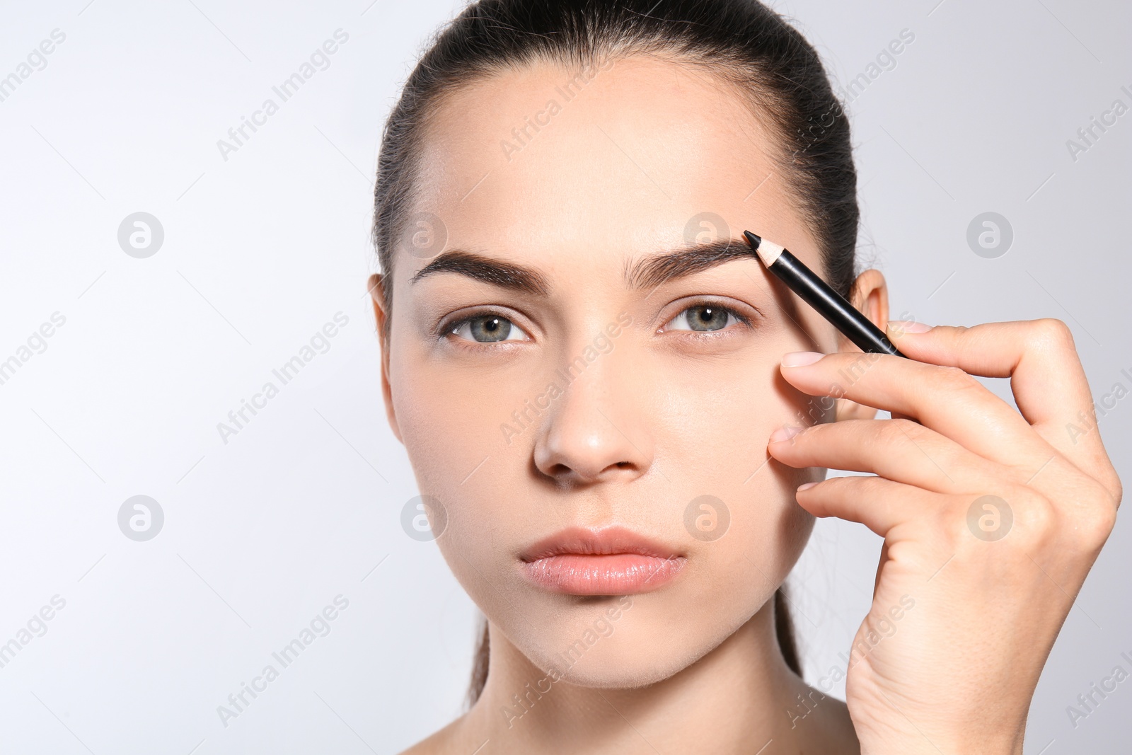 Photo of Young woman correcting shape of eyebrow with pencil on light background