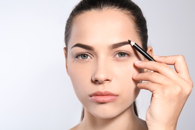 Photo of Young woman correcting shape of eyebrow with pencil on light background