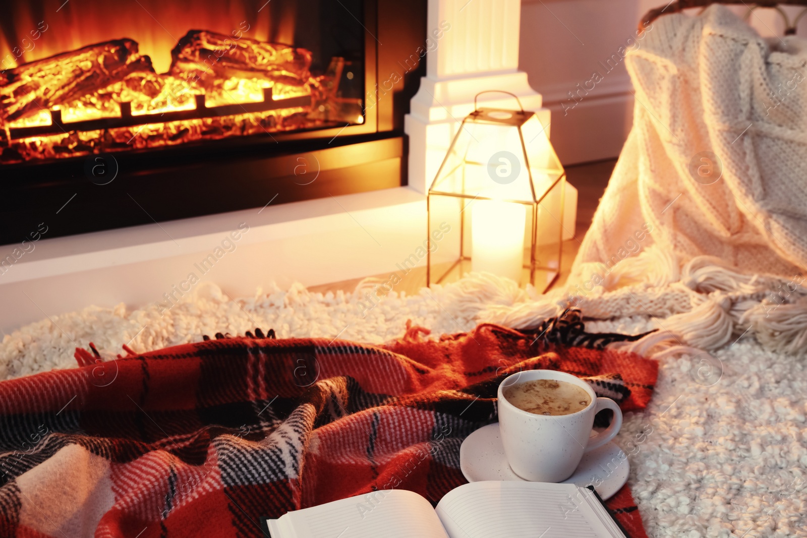 Photo of Cup of hot drink and book on blanket near fireplace indoors. Magic atmosphere