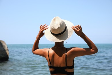 Young woman in stylish bikini and hat near sea, back view