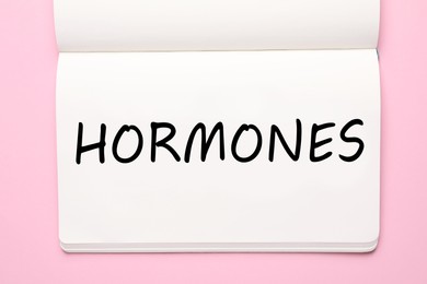Photo of Notebook with word Hormones on pink background, top view