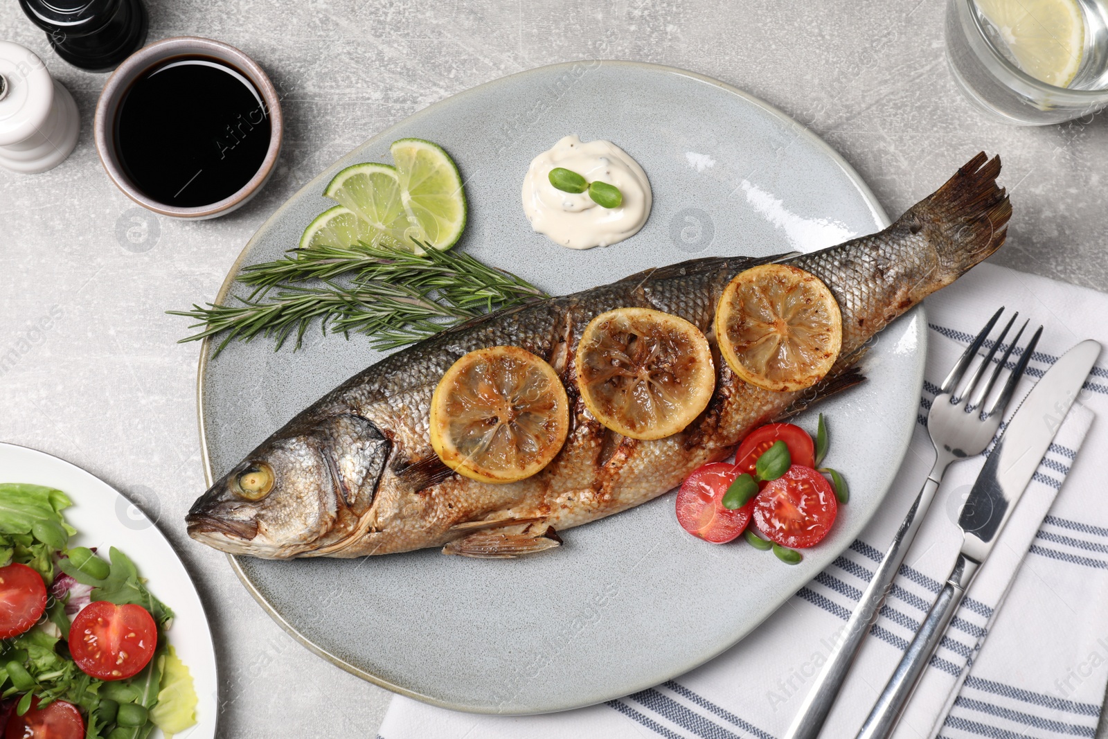 Photo of Delicious roasted sea bass fish served with lemon, rosemary and sauce on light grey table, flat lay