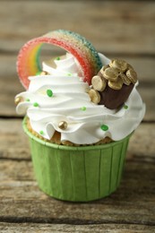 Photo of St. Patrick's day party. Tasty cupcake with sour rainbow belt and pot of gold toppers on wooden table, closeup