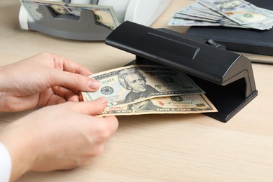 Woman checking dollar banknotes with currency detector at wooden table, closeup. Money examination device