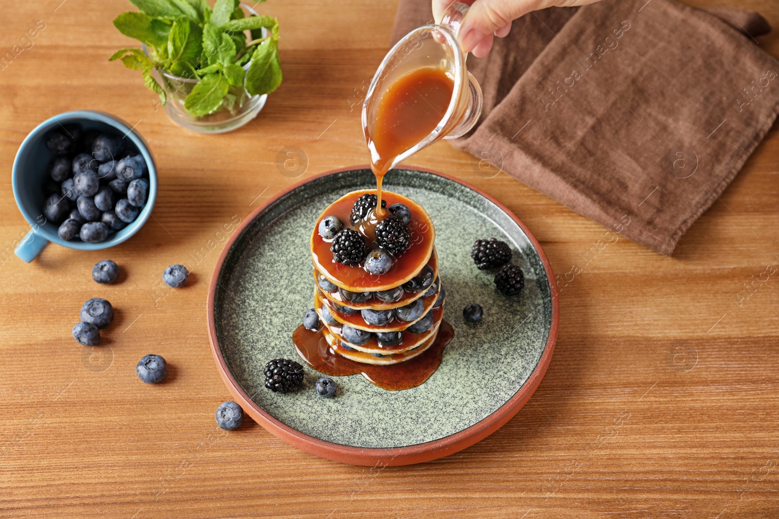 Photo of Woman pouring syrup onto tasty pancakes with berries on plate, closeup