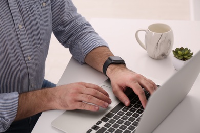 Young man with smart watch working on laptop at table in office, closeup