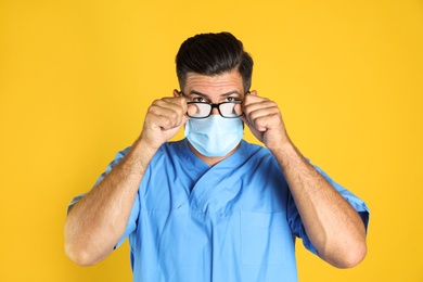 Doctor wiping foggy glasses caused by wearing medical mask on yellow background