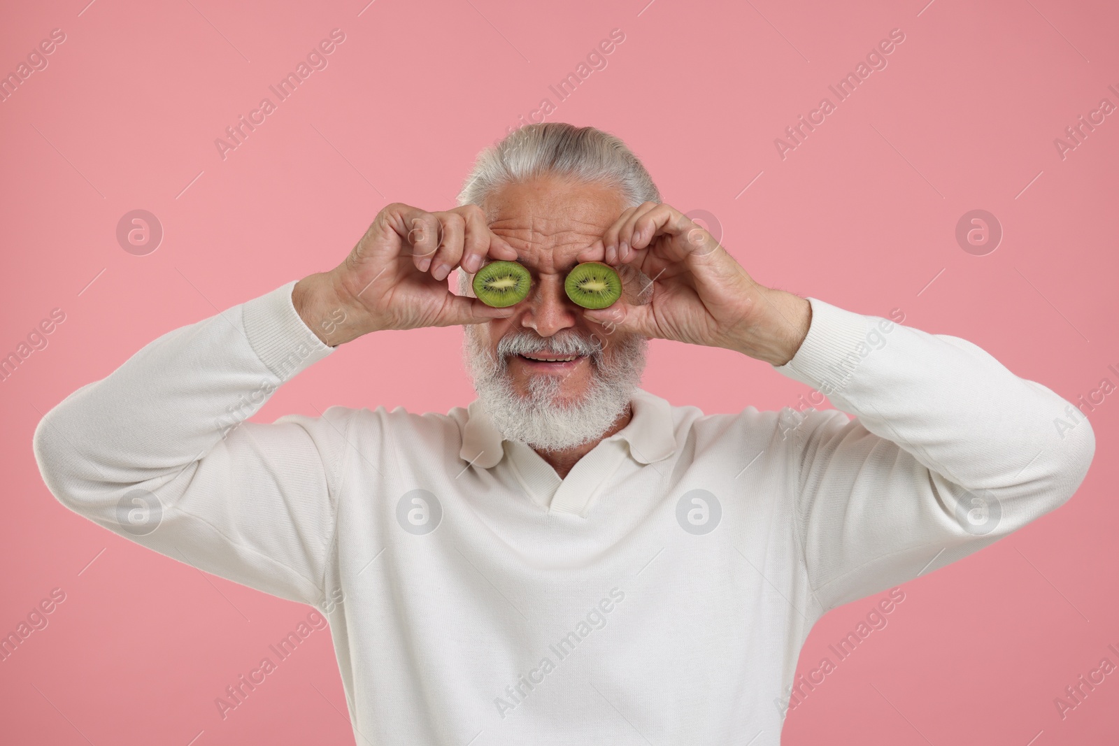 Photo of Senior man covering eyes with halves of kiwi on pink background