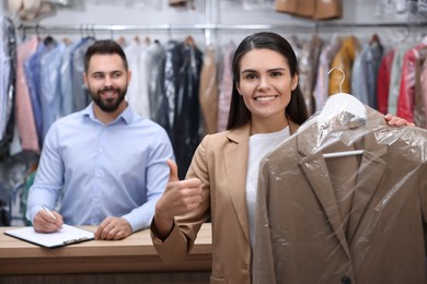 Photo of Dry-cleaning service. Happy woman holding hanger with coat and showing thumb up indoors. Worker taking notes at workplace