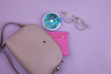 Stylish women's bag with plastic pill box, glasses and stationery on lilac background, flat lay