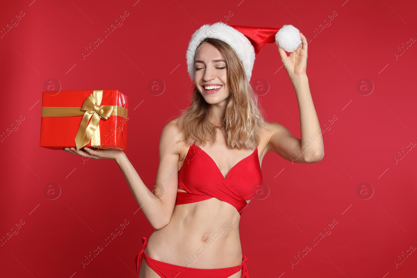 Photo of Sexy young woman in bikini and Santa hat with gift box on red background. Christmas celebration