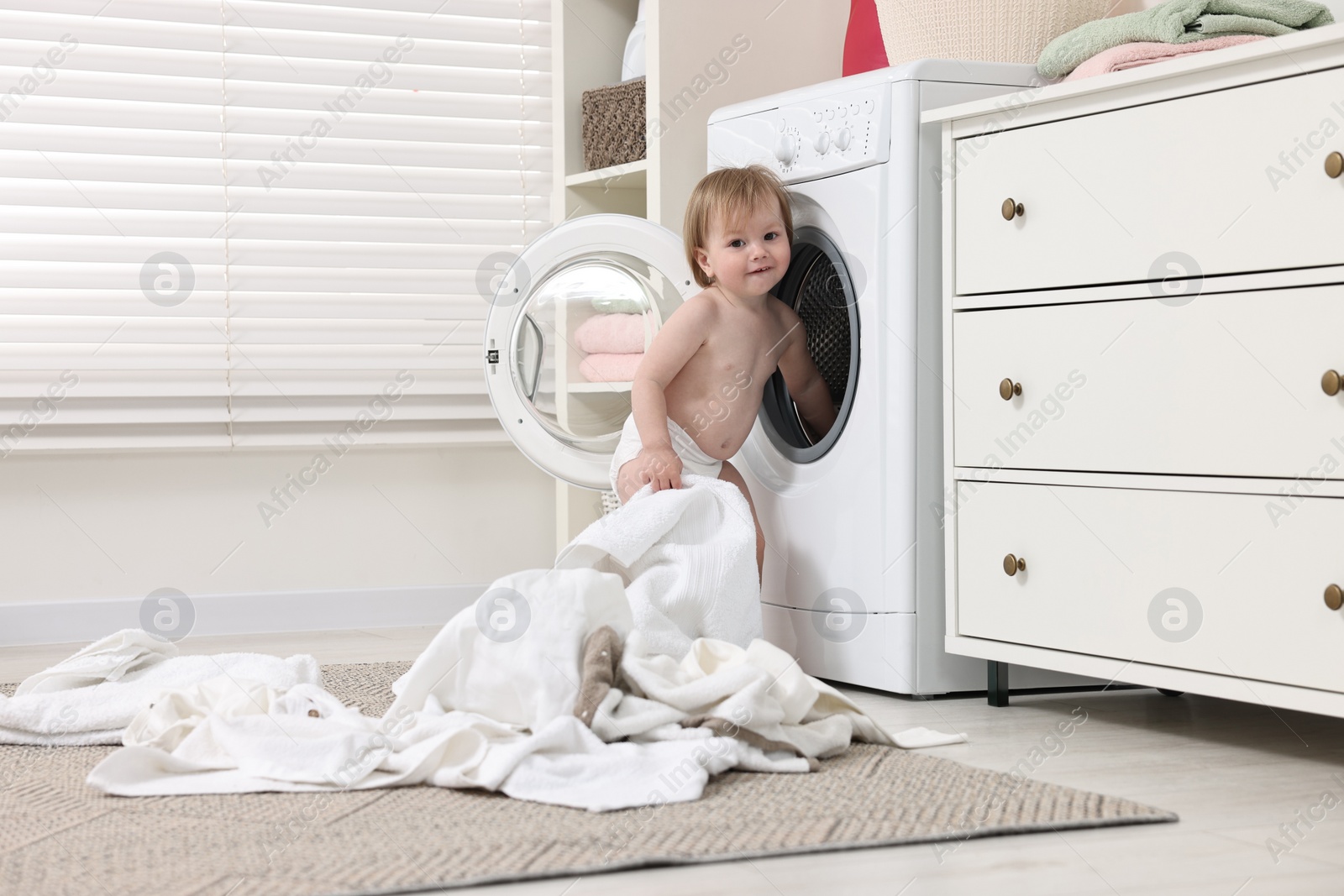 Photo of Little girl pulling baby clothes out of washing machine indoors