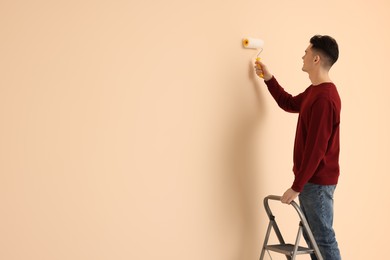 Young man painting wall with roller on stepladder indoors, space for text. Room renovation