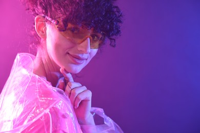 Photo of Beautiful young woman in transparent coat and sunglasses posing on color background in neon lights