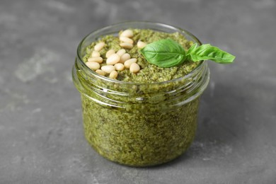 Photo of Jar with delicious pesto sauce, pine nuts and basil leaves on grey table, closeup