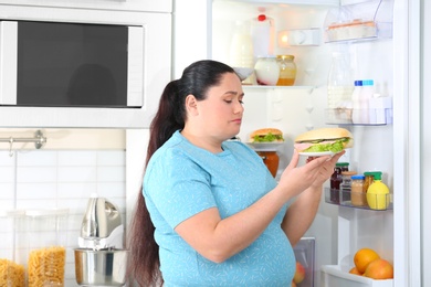 Sad overweight woman taking sandwich from refrigerator in kitchen. Failed diet