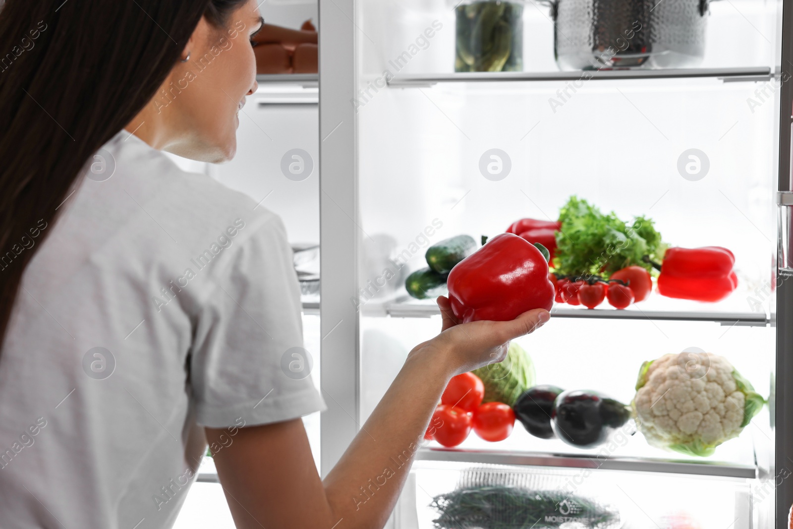 Photo of Young woman taking red bell pepper out of refrigerator at night, closeup