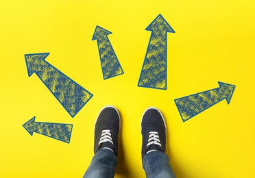 Image of Choosing future profession. Teenager standing in front of drawn signs on yellow background, top view. Arrows pointing in different directions symbolizing diversity of opportunities