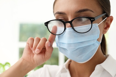 Photo of Woman wiping foggy glasses caused by wearing medical mask indoors, closeup