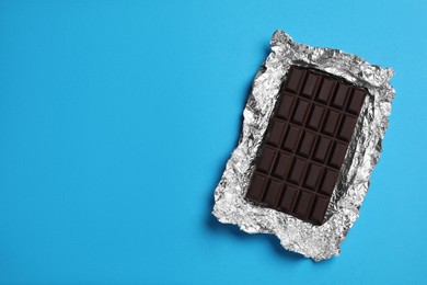 Photo of Delicious dark chocolate bar with foil on light blue background, top view. Space for text