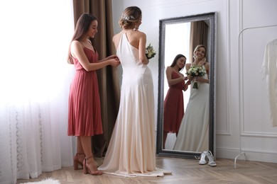 Photo of Gorgeous bride in beautiful wedding dress and her friend near mirror in room