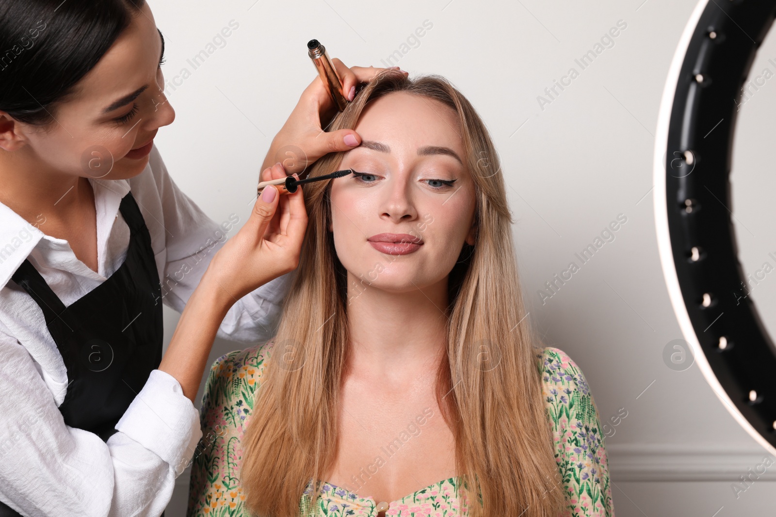 Photo of Professional makeup artist working with beautiful young woman near white wall indoors. Using ring lamp