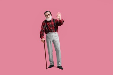 Senior man with walking cane waving on pink background