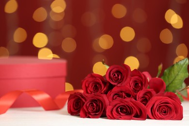 Photo of Beautiful red roses and gift box on white table against blurred lights, space for text. St. Valentine's day celebration