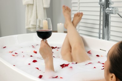 Woman with glass of wine taking bath in tub with foam and rose petals indoors, closeup