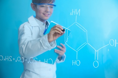 Schoolboy writing chemistry formula on glass board against color background. Space for text