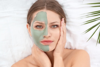 Beautiful woman with clay facial mask and tropical leaf on white fabric, above view