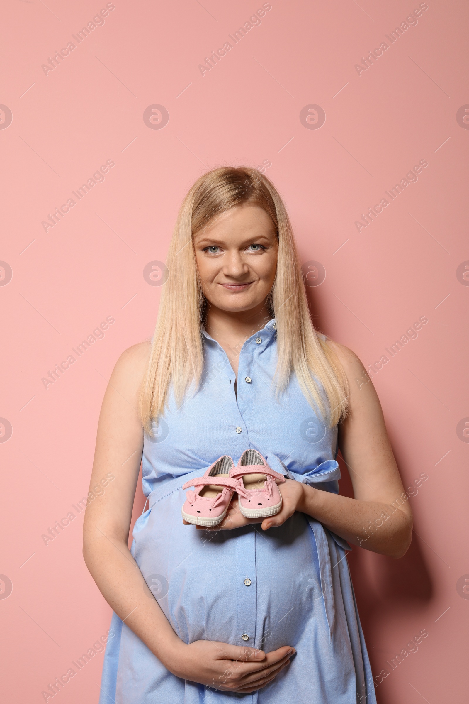 Photo of Beautiful pregnant woman in dress holding baby booties near tummy on color background