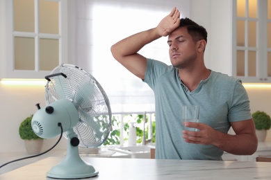 Man with glass of water and modern fan at table in kitchen. Summer heat