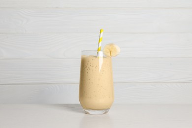 Glass of tasty smoothie with banana piece and straw on white wooden table