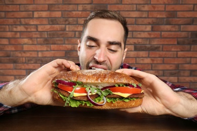 Photo of Young hungry man eating tasty sandwich on brick wall background