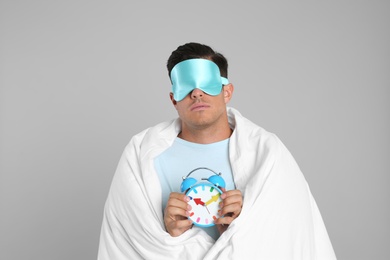 Man in sleeping mask wrapped with blanket holding alarm clock on grey background