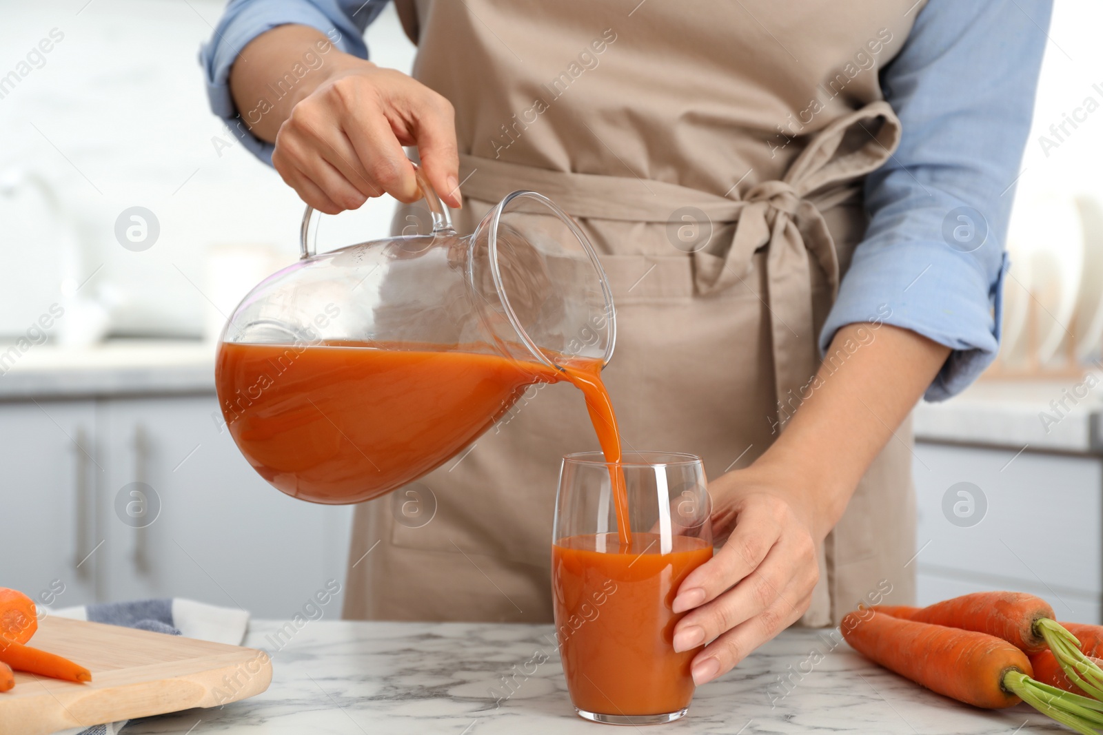 Photo of Woman pouring tasty carrot juice from jug into glass at table indoors, closeup