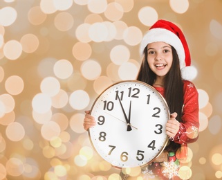 New Year countdown. Cute little girl in Santa hat holding clock against blurred lights on background