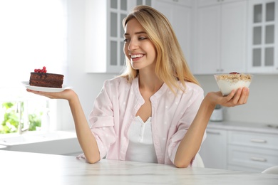 Photo of Woman choosing between yogurt with granola and cake at white table in kitchen