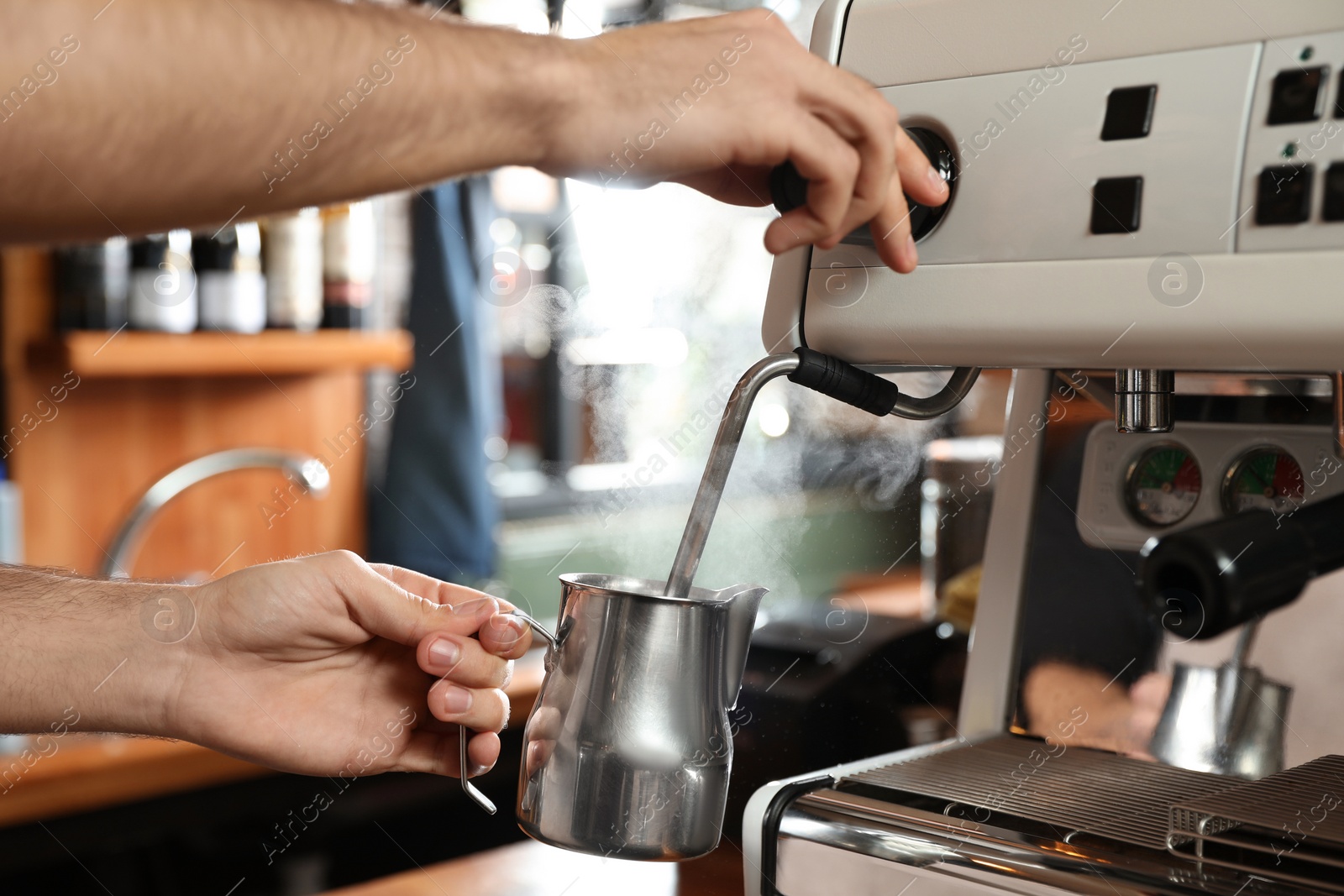 Photo of Barista steaming milk in metal jug with coffee machine wand at bar counter, closeup