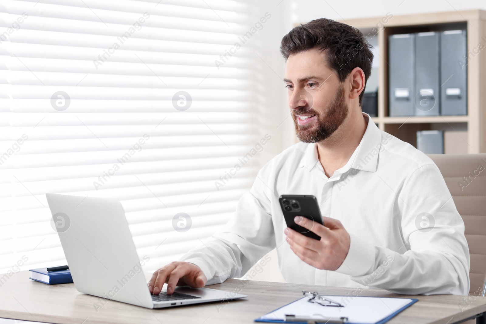 Photo of Smiling man with smartphone using laptop at table in office