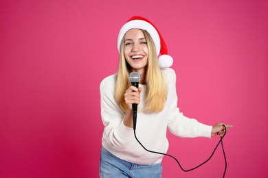 Happy woman in Santa Claus hat singing with microphone on pink background. Christmas music