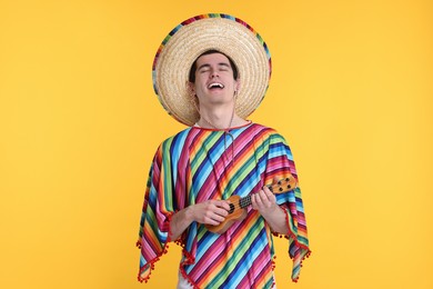 Photo of Young man in Mexican sombrero hat and poncho playing ukulele on yellow background